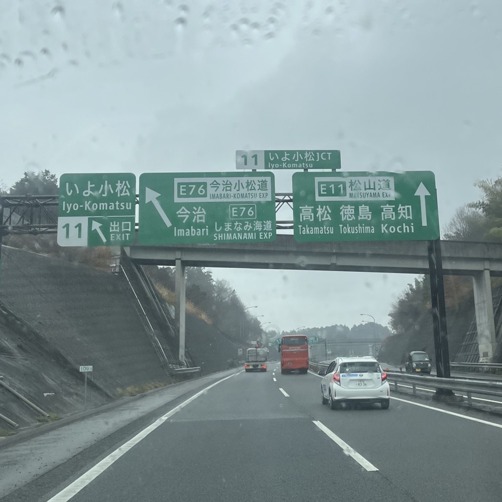 雨の中の松山自動車道、広い道の上には大きな緑の各方面の標識看板が上がっている