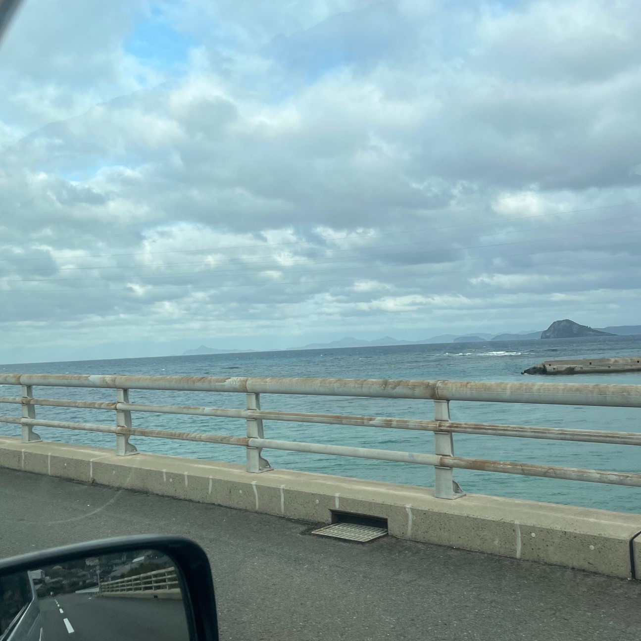 島の海沿いの道、広い海には小島が見え雲がかかった空が広がっている