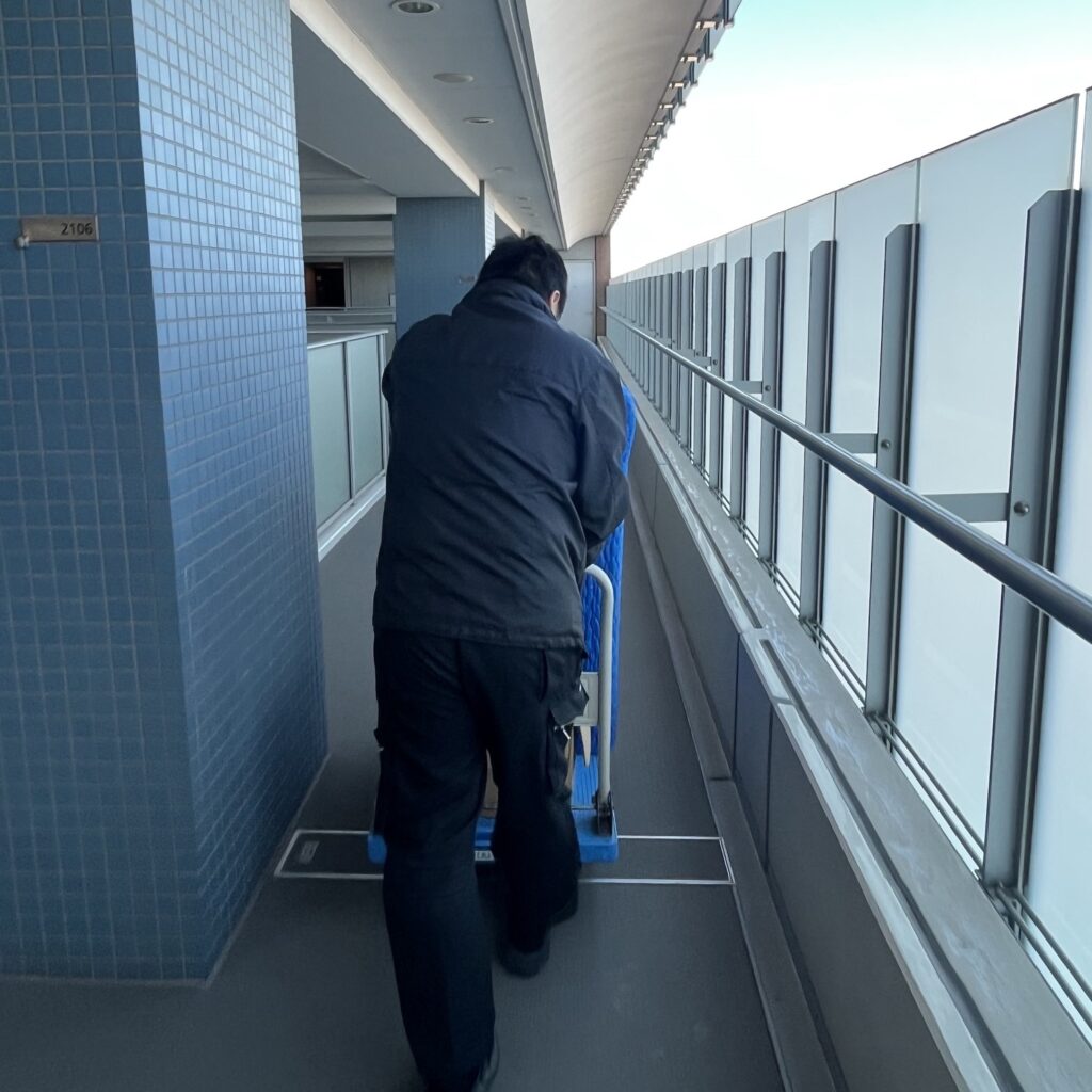 マンション上層階の風が吹き抜ける通路でも職人が丁寧にお仏壇を運んでいる